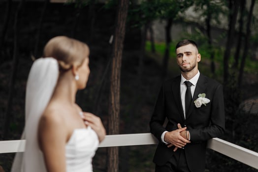 A bride and groom are standing on a balcony, with the groom looking at the bride. The bride is wearing a white dress and a veil, while the groom is wearing a black suit and tie photo