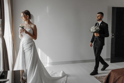A bride and groom are standing in a room with a window. The bride is holding a bouquet and the groom is holding a bouquet as well photo