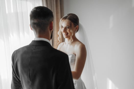 A bride and groom are standing in front of a window, smiling at each other. The bride is wearing a white dress and the groom is wearing a suit. Scene is happy and joyful photo