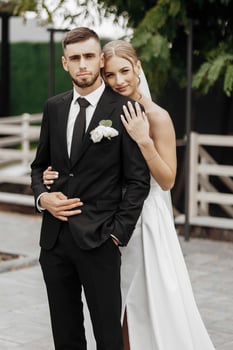 A bride and groom are posing for a picture in front of a fence. The bride is wearing a white dress and the groom is wearing a black suit. Scene is happy and celebratory photo