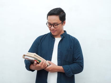 Young Asian man holding books smiling happy and positive while reading a book. Asian student flipping a book page over white background photo