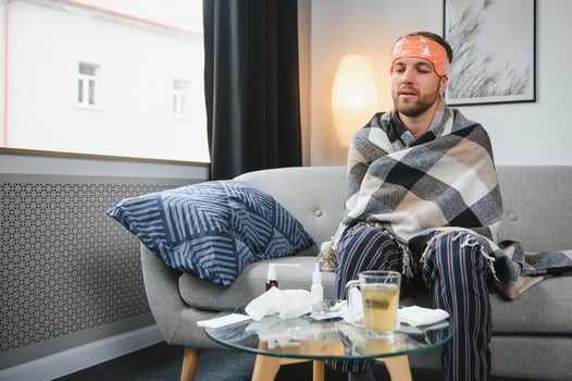 Sick bearded man who has bad cold or seasonal flu sitting on couch at home. Guy with fever wearing warm plaid shivering with worried face expression. photo