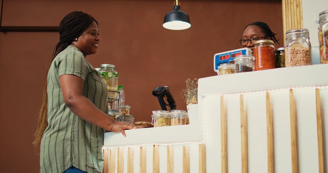 Employee putting jars of bulk items on scale at checkout, helping regular customer with choosing groceries from local farmers market. African american vendor selling organic goods. Handheld shot. photo