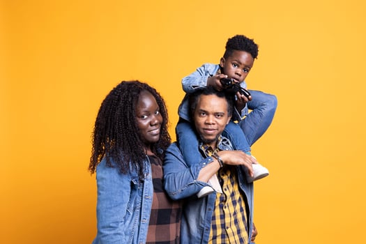 African american dad carrying his toddler on his shoulders against background, sweet kid having fun on camera with a joystick in his hand. Joyful young mother and father bonding with their son. photo
