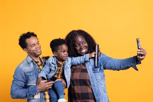 Happy african american family of three taking photos in studio, posing together with their infant baby boy. Cheerful mother and father takes selfies on smartphone, making memories.