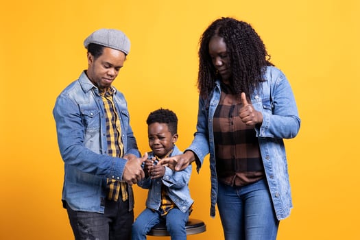 Smiling parents showing thumbs up gesture on camera, posing next to their young child sitting on the chair. Happy african american parents giving like sign, approving something in studio. photo