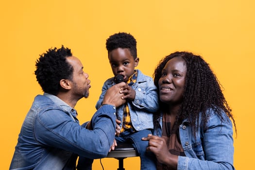Cheerful adorable small child singing a song on microphone in studio, parents smiling and dancing on his music. Proud mother and father having fun with their happy son on camera. photo