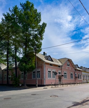 A picturesque pink wooden house sits gracefully at a sunny street corner, framed by tall trees and a clear blue sky, embodying a quaint and peaceful charm. photo