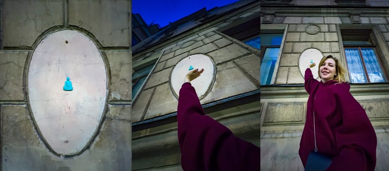 A cheerful woman in a maroon sweater reaches up towards a small blue Buddha figurine attached to the wall of a historic building. The playful interaction with the unique street art adds a touch of whimsy to the urban scene photo
