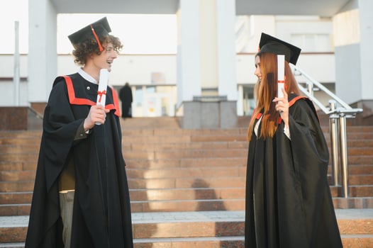 Portrait of happy graduates. Two friends in graduation caps and gowns standing outside university building with other students in background, holding diploma scrolls, and smiling photo