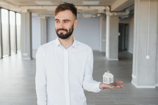 Indoor portrait of happy male home owner showing key to his new house. Joyful handsome single young Caucasian man in casual shirt standing in room, holding keys and smiling. Real estate concept photo