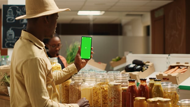 Business owner presenting blank greenscreen display on phone, showing isolated mockup layout in zero waste eco store. African american vendor using chromakey template on mobile phone. Camera 1. photo