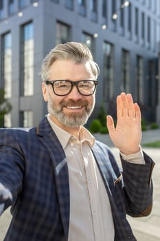 Cheerful senior male executive with glasses greeting with a hand wave, standing outside a modern office building in a suit. photo