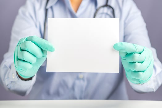 Close-up of doctor hands holding a blank white paper while sitting on a chair with gray background in the hospital or clinic. Space for text. Medical concept photo