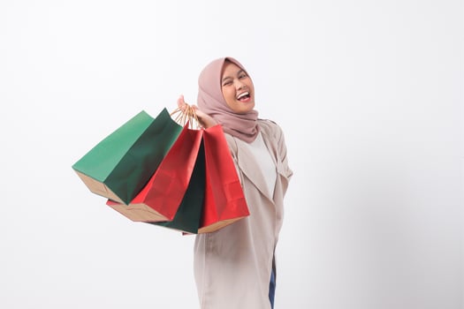 Portrait of excited Asian hijab woman in casual suit holding and showing colorful paper shopping bags. Shopaholic girl and discount buying concept. Isolated image on white background photo