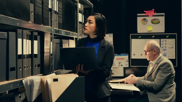 Detective evaluating old archived folders to organize all cases based on crimes, digitizes documents to preserve evidence on laptop. Private agent searching for forensics notes. Camera B. photo
