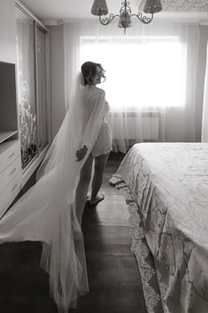 A woman in a white gown stands in front of a bed. The room is dimly lit, and the woman is waiting for someone photo