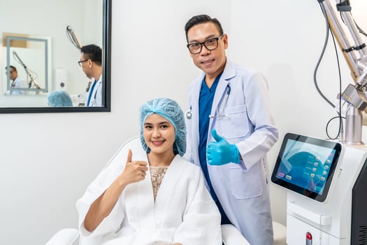 Front view of asian male cosmetologist and young woman patient with blue cap on head sitting in chair in cosmetological clinic , smiling, looking at camera, Concept of cosmetology. photo