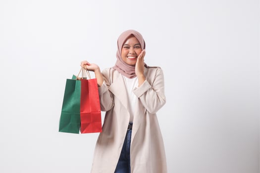 Portrait of shocked Asian hijab woman in casual suit holding paper shopping bags while covering mouth. Shopaholic girl and discount buying concept. Isolated image on white background photo