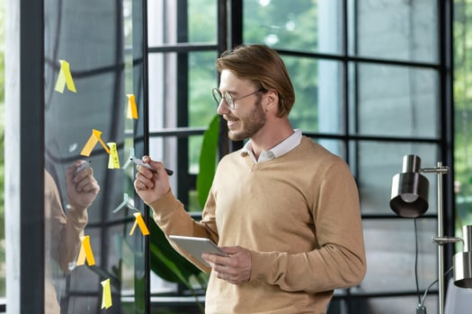 Happy man in casual clothes standing near a glass note board and sticking colorful stickers with reminders and plans on it, writing something with a pen, office work planning concept. photo