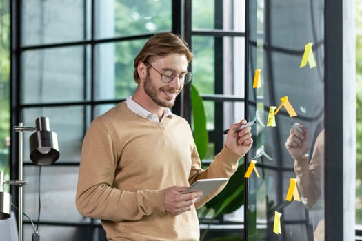 Smiling young man businessman, programmer, freelancer standing in the office, looking at a tablet, drawing a progress graph on a transparent board. Glue colored papers, notes, stickers, hints. photo