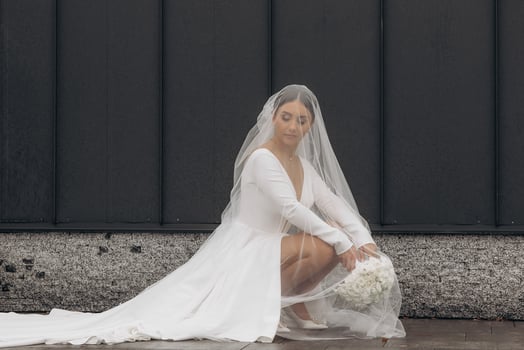 A woman in a white wedding dress is kneeling down and holding a bouquet of flowers. She is wearing a veil and is surrounded by a black background. Concept of elegance and romance photo