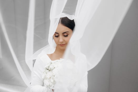 A woman is wearing a white dress and holding a bouquet of flowers. She is standing in front of a white curtain, which is partially covering her. Concept of intimacy and romance photo