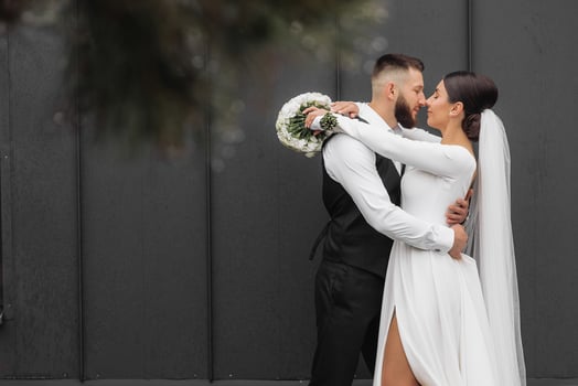 A bride and groom are embracing each other in front of a black wall. The bride is wearing a white dress and the groom is wearing a suit. The couple is holding a bouquet and a small vase photo