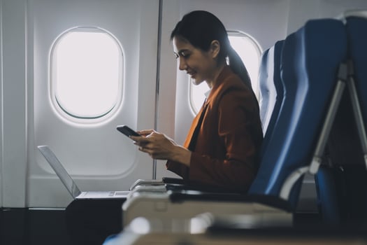 Asian female tourist checks notifications on smartphone while seated on airplane A businesswoman happily shares media from her phone, taking photos of the view from the window during a flight.