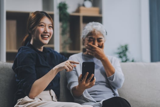 Cheerful mother and pretty teenager kid relaxing at cozy home, using mobile phone for Internet communication, resting on sofa with digital gadget, enjoying leisure, looking at camera photo