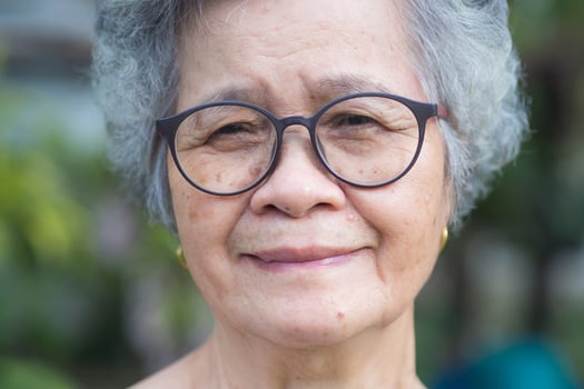 Portrait of an elderly woman with short gray hair, wearing glasses, smiling, and looking at the camera while standing in a garden. Aged people and relaxation concept photo