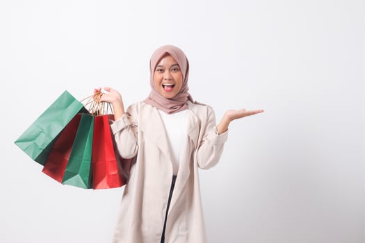 Portrait of excited Asian hijab woman in casual suit holding paper shopping bags while pointing with finger. Shopaholic girl and discount buying concept. Isolated image on white background photo