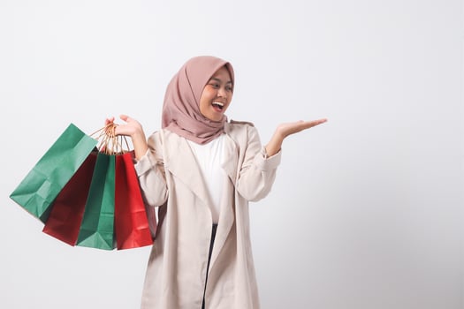 Portrait of excited Asian hijab woman in casual suit holding paper shopping bags while pointing with finger. Shopaholic girl and discount buying concept. Isolated image on white background photo