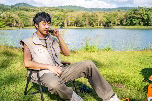 Young Asian traveling man relax and enjoy outdoor lifestyle camping in forest mountain on summer holiday travel vacation. Attractive male drinking coffee while enjoys drink among green trees photo