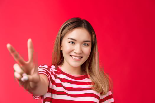 Optimsitic positive young asian female, support lgbt community, pull hand forward show peace victory sign, smiling happily feel lucky, stand red background joyful, enjoy summer vacation photo