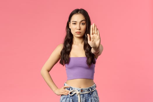 Waist-up portrait of confident stylish young woman stand in no trespassing pose, raise hand in stop sign, look camera skeptical, disagree, dont permit step closer, warning, pink background photo