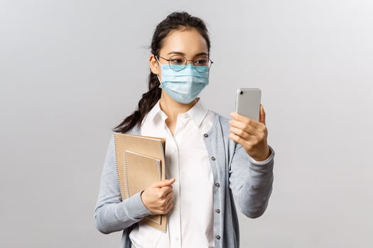 Covid19, virus, health and medicine concept. Young asian girl in medical mask, holding notebooks and work reports, taking selfie on smartphone, videocalling friends while social distancing photo