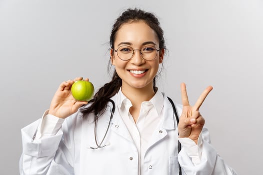 Portrait of enthusiastic young happy smiling female doctor in white coat, show peace sign and green apple, asking people stay healthy, eat more fruits and vitamins, grey background photo