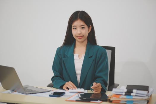 Asian businesswoman in red suit calculating company sales with calculator, laptop, and tablet on table Interior of businesswoman office at the modern workstation. photo