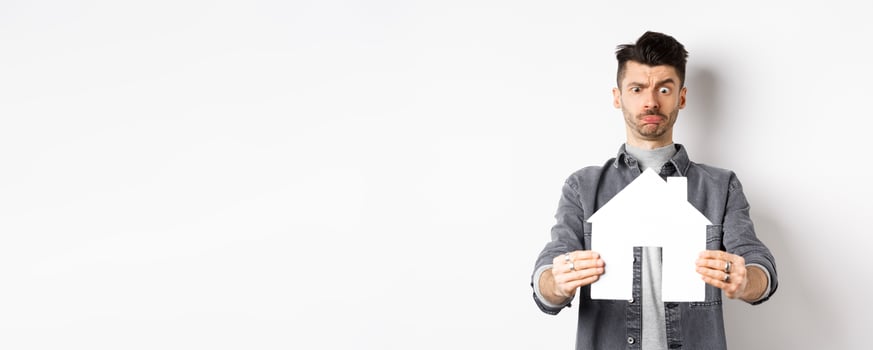 Real estate and insurance concept. Confused guy look at paper house cutout unsure, frowning perplexed, standing on white background photo