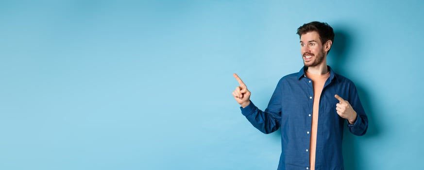 Smiling handsome guy pointing and looking left at empty space, showing logo, standing on blue background photo