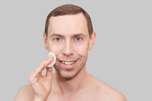 Man doing home skin care routine. Cotton pad near male chin. Dermatology treatment. Cream facial apply. Cosmetology guy portrait. Cleaning lotion. Skincare hygiene. Copyspace. Isolated on grey photo