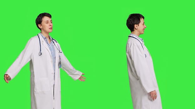 Friendly medic waving hello and greeting patients against greenscreen backdrop, smiling before her exam appointments. Woman physician with white coat welcoming people and being happy. Camera B. photo