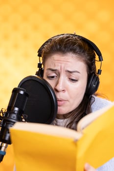 Woman frowns while doing voiceover reading of book to produce audiobook using melodramatic acting. Narrator glowers, using storytelling skills to portray character, recording novel, studio background photo