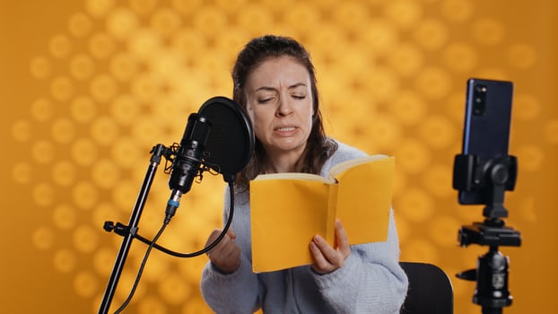 Online star films vlog at work using smartphone on tripod, passionately reading book to record audiobook. Narrator doing content creation with phone, producing digital recording of novel, camera B photo