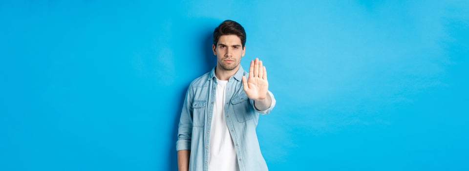 Serious man frowning and saying no, extending hand to shop stop sign, prohibit action, standing against blue background photo