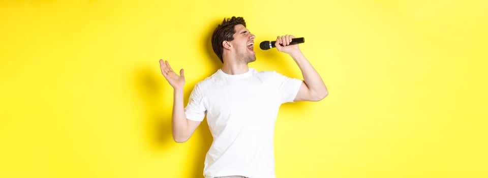 Young man singer holding microphone, reaching high note and singing karaoke, standing over yellow background photo