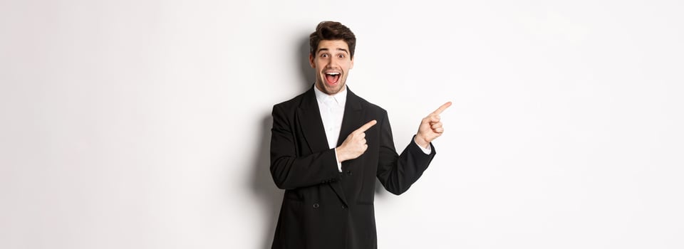 Portrait of excited and happy man in suit showing advertisement, pointing fingers at upper left corner and smiling amazed, standing over white background photo