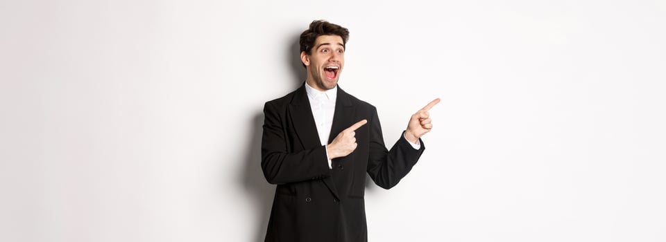 Portrait of amazed and happy handsome man in black suit, pointing and looking left impressed, showing awesome christmas promo, standing over white background photo