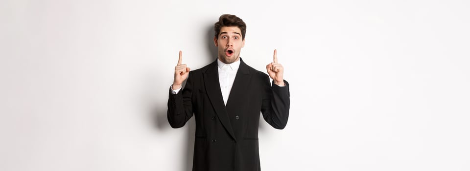 Portrait of surprised handsome businessman in black suit, saying wow and looking amazed, pointing fingers up at copy space, standing over white background photo